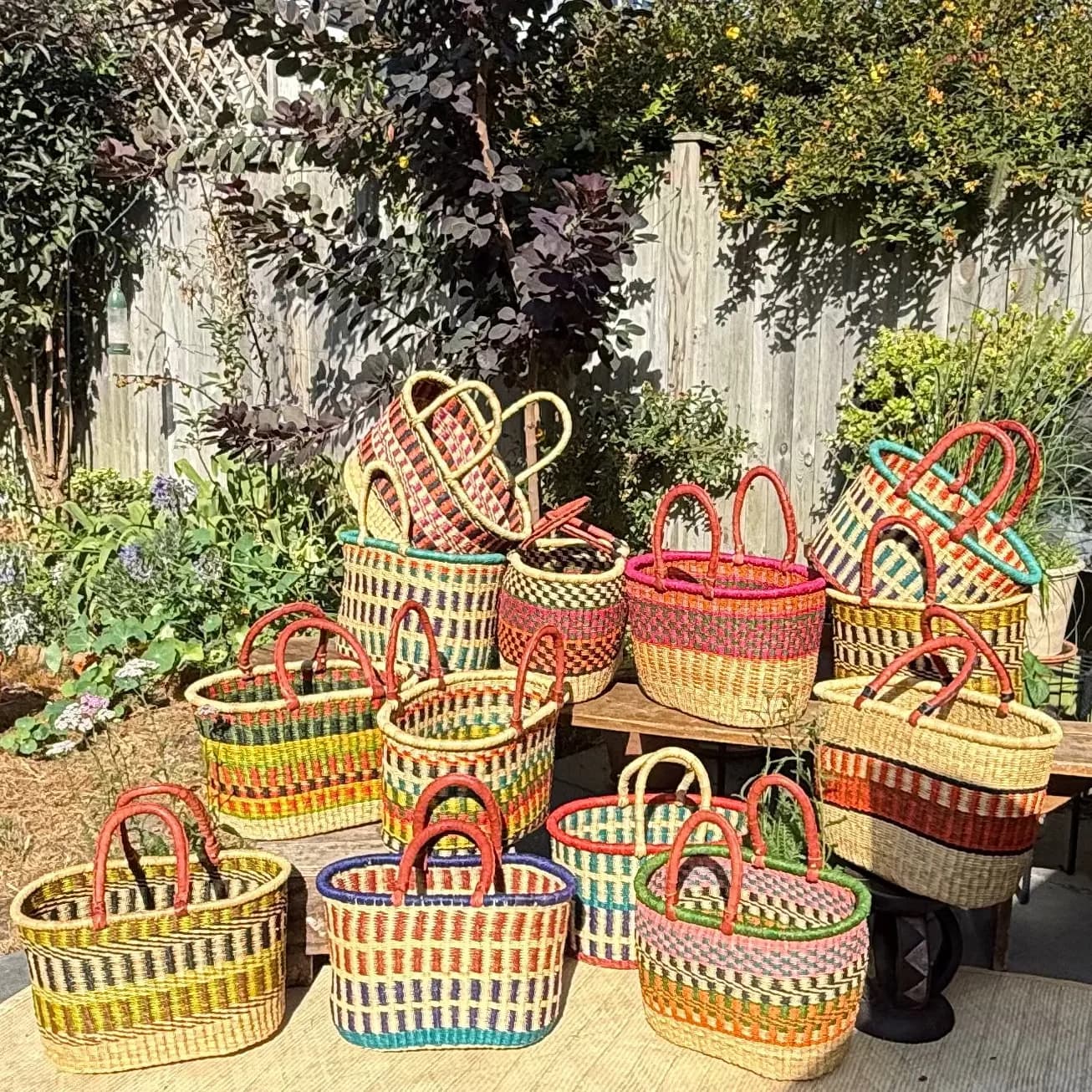 A collection of colorful woven baskets and wicker decor displayed on a patio in a backyard setting with a wooden fence, greenery, and a hanging textile.