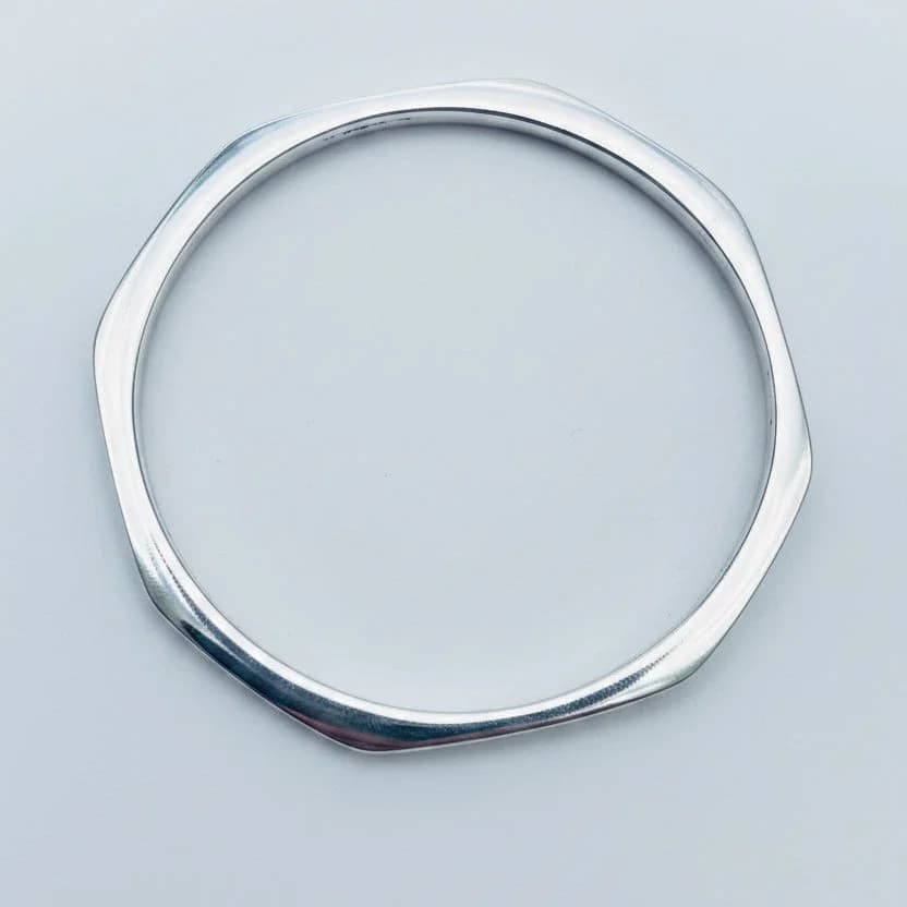 A polished silver-toned metal bangle bracelet with a wavy, organic hexagonal outline, displayed on a solid light blue background.