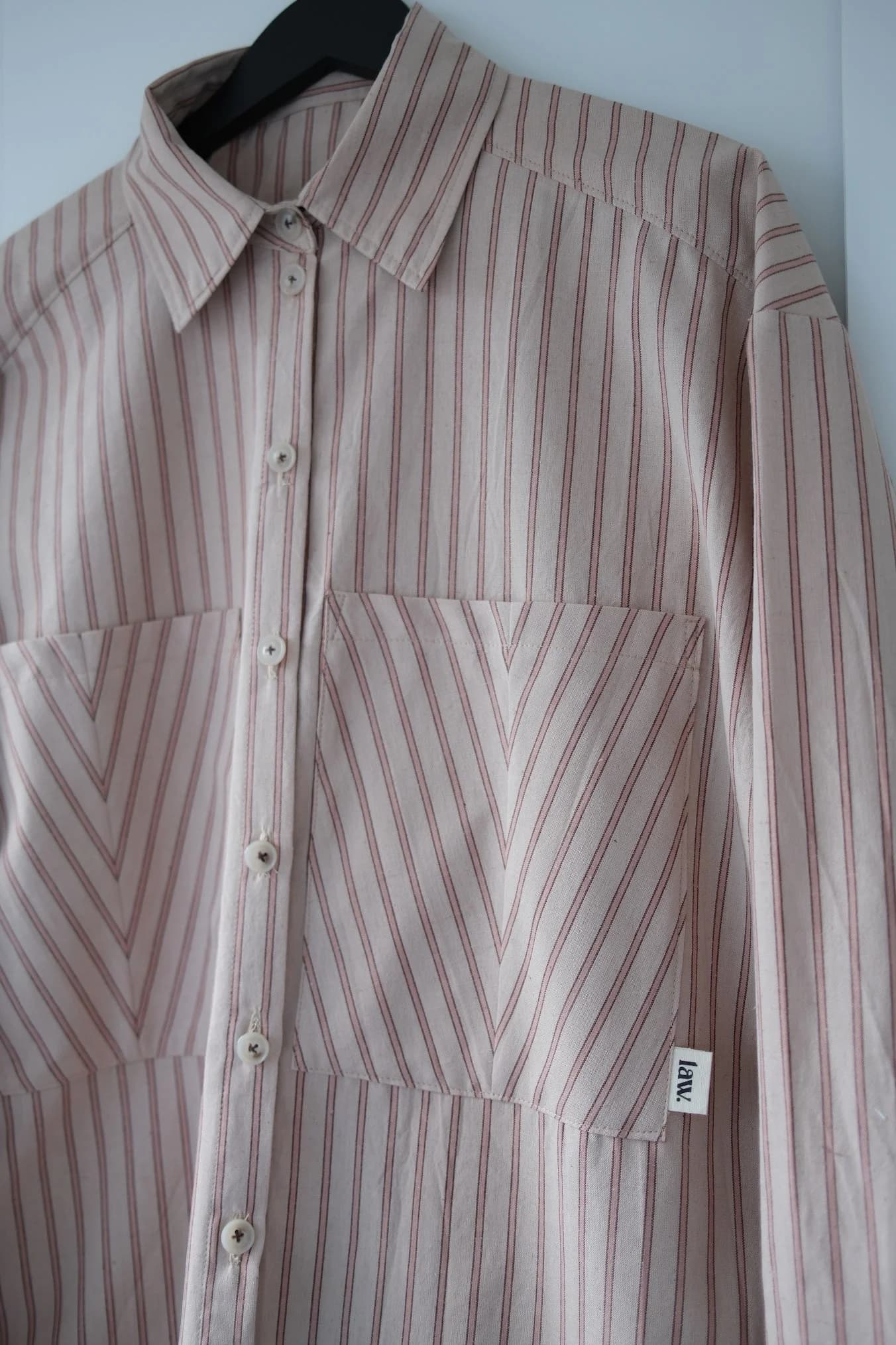 A long-sleeve button-up shirt with vertical and chevron stripe patterns in beige and brown, hanging on a black hanger against a light gray wall.