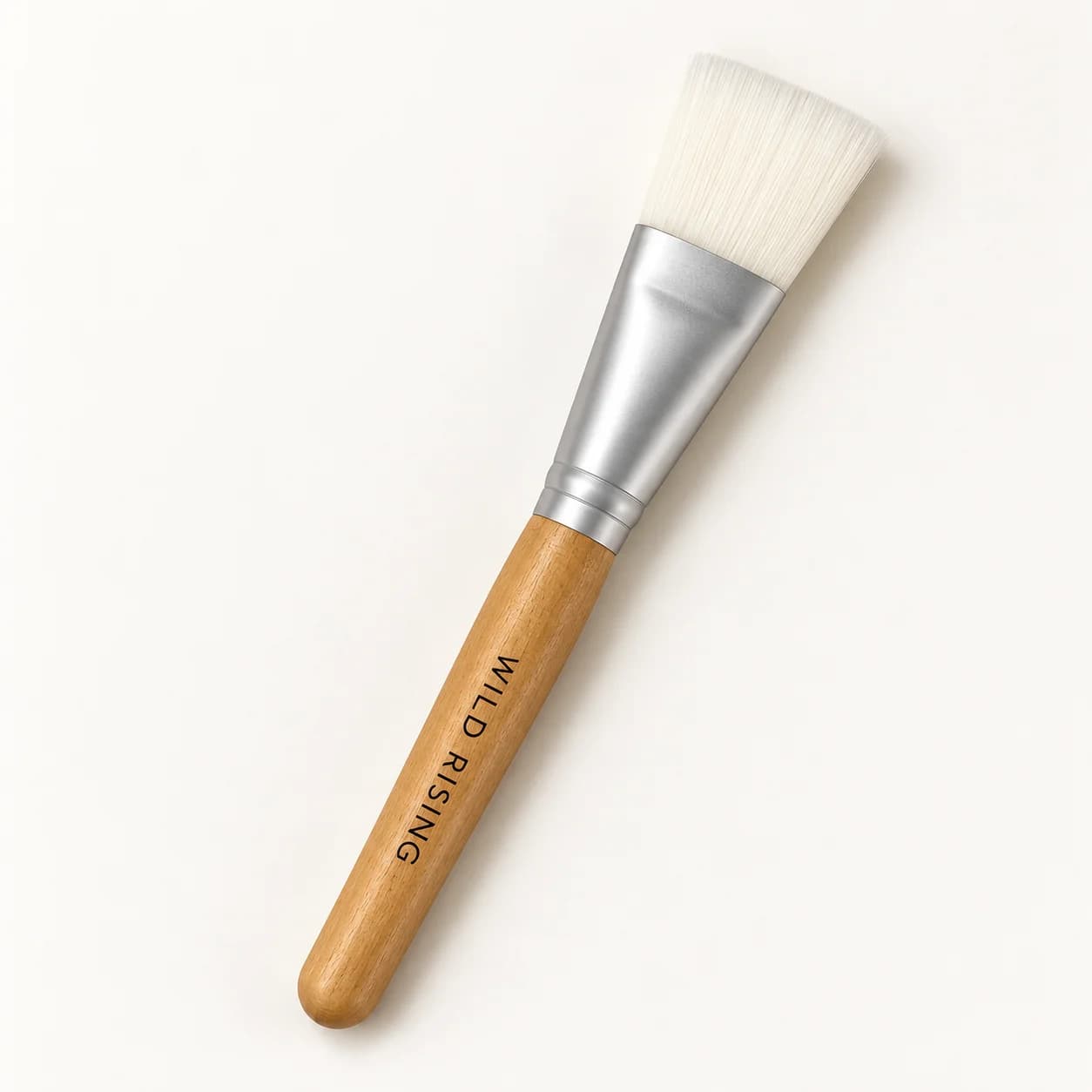A flat makeup brush with white bristles, a silver metal ferrule, and a light wood handle. The handle is printed with the brand name 'WILD RISING'.