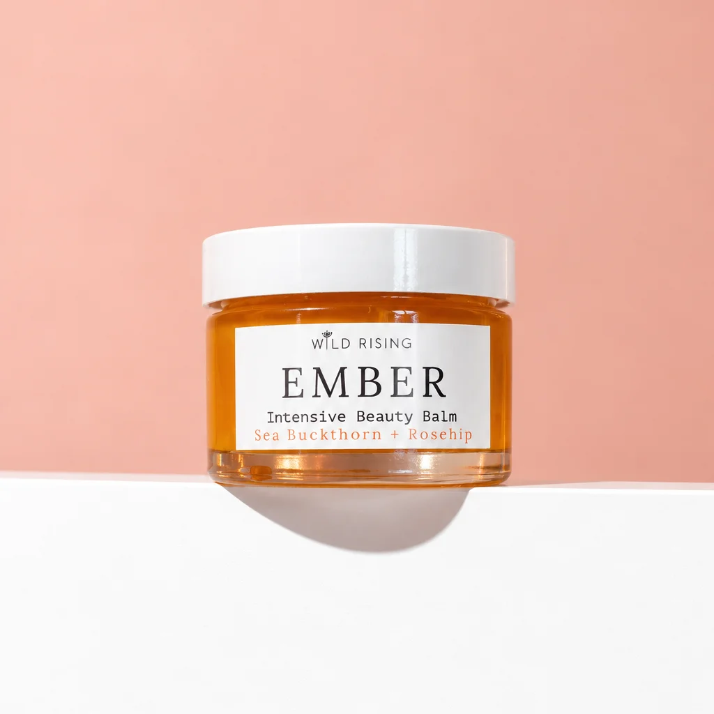A jar of Wild Rising Ember Intensive Beauty Balm with amber-colored balm visible through a clear jar and a white lid, set against a split peach and white background.