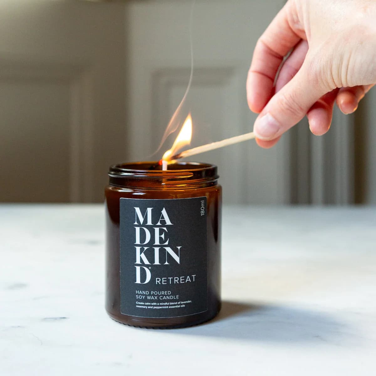 MadeKind soy wax candle in an amber glass jar candle. Lit with a wooden match held by a person's hand, set on a light marble surface. Scented with essential oils to make your home smell like a retreat.