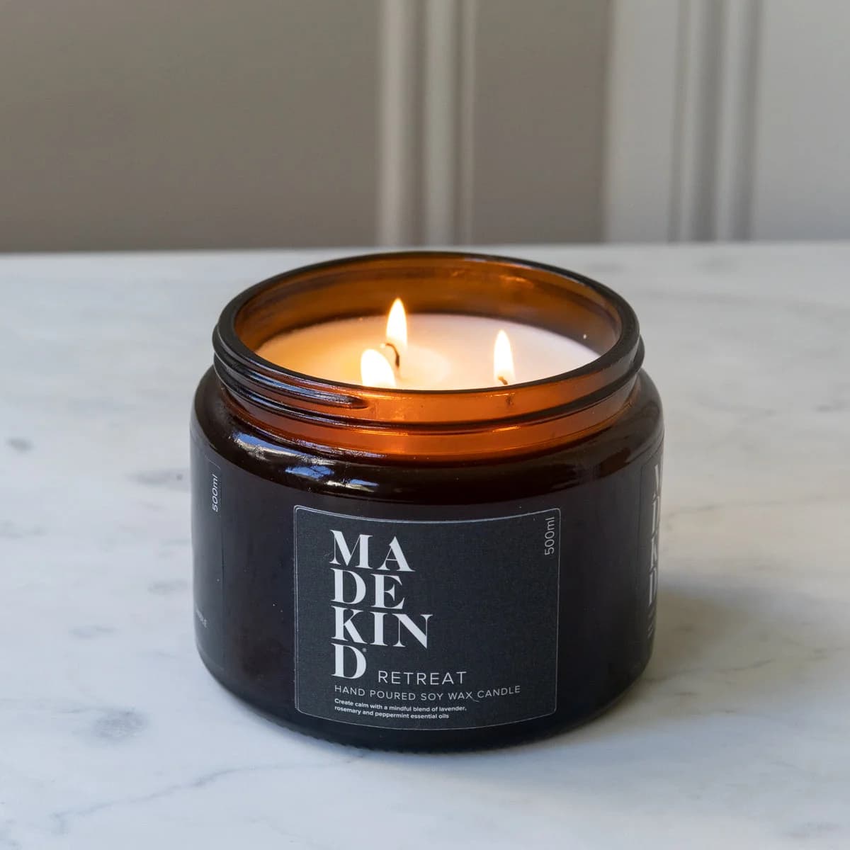 A lit amber glass jar MadeKind soy wax candle on a white marble surface with a neutral indoor background. Scented with Retreat essential oils.