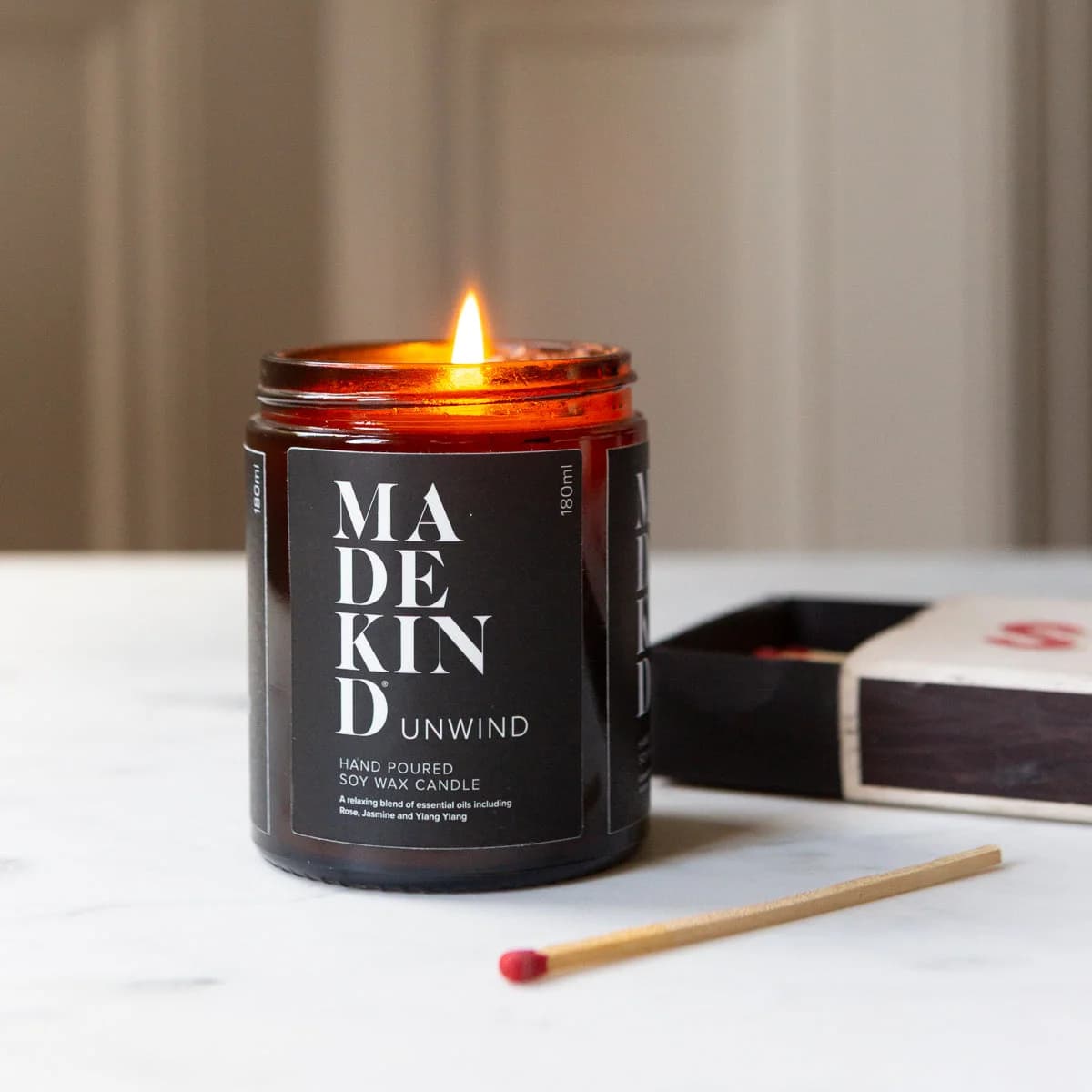 A lit amber glass jar MadeKind candle with a black label reading MADE KIND UNWIND, set on a light marble surface with a match in front and an open box behind it.