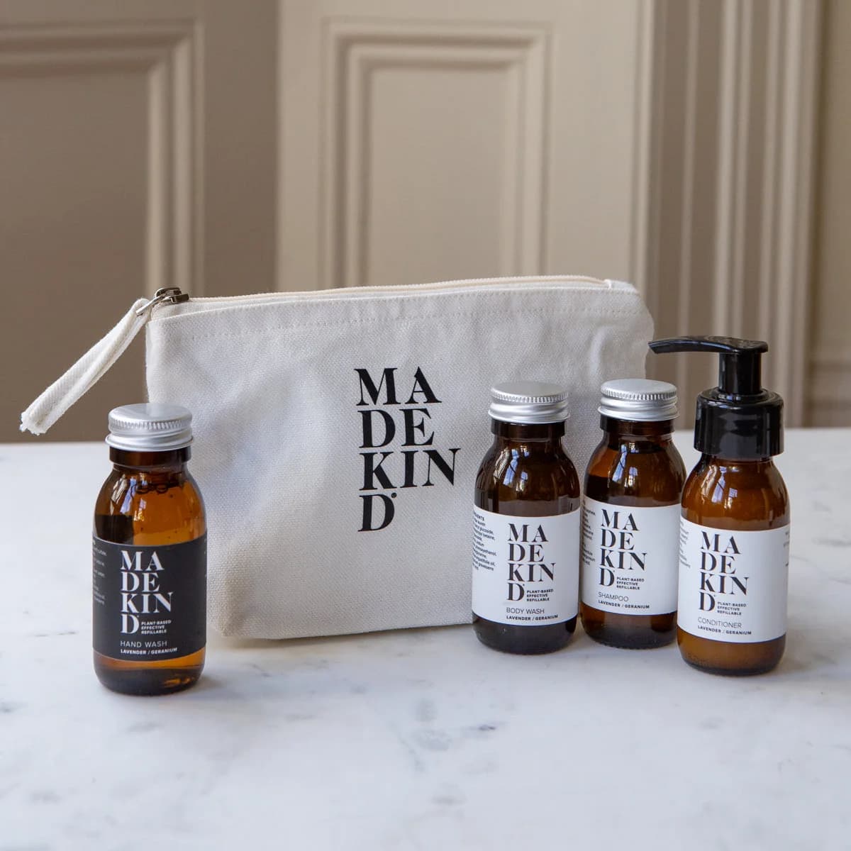 Toiletries set arranged on a white marble surface in front of a cream canvas pouch. The set includes four amber glass bottles and a pump bottle with white and black labels reading MADE KIND, plus a cream zip pouch with black MADE KIND branding.