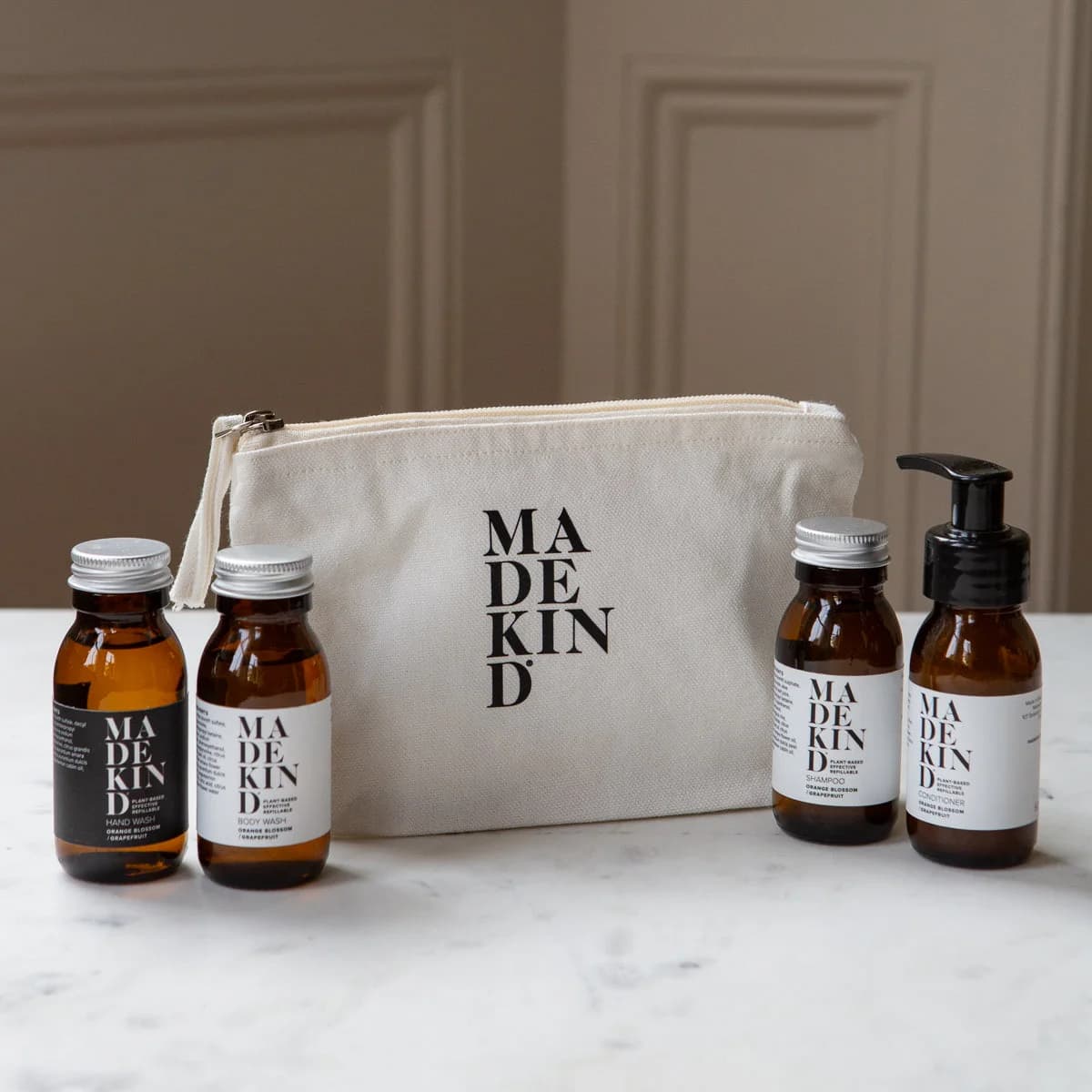 A set of MadeKind mini cleaning products arranged on a white marble surface in front of a beige canvas pouch. The set includes four amber glass bottles with cleaning products in, with white labels and metal caps, plus a cream canvas zip pouch with black text.