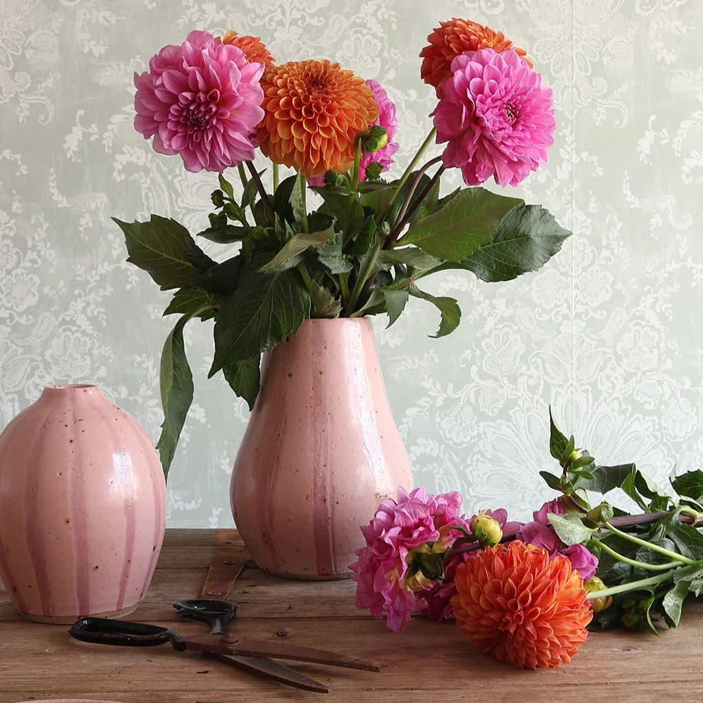 A pink ceramic vase filled with bright pink and orange dahlias sits on a rustic wooden table, alongside a matching pink vase, scissors, and additional flowers against a pale patterned wallpaper background.