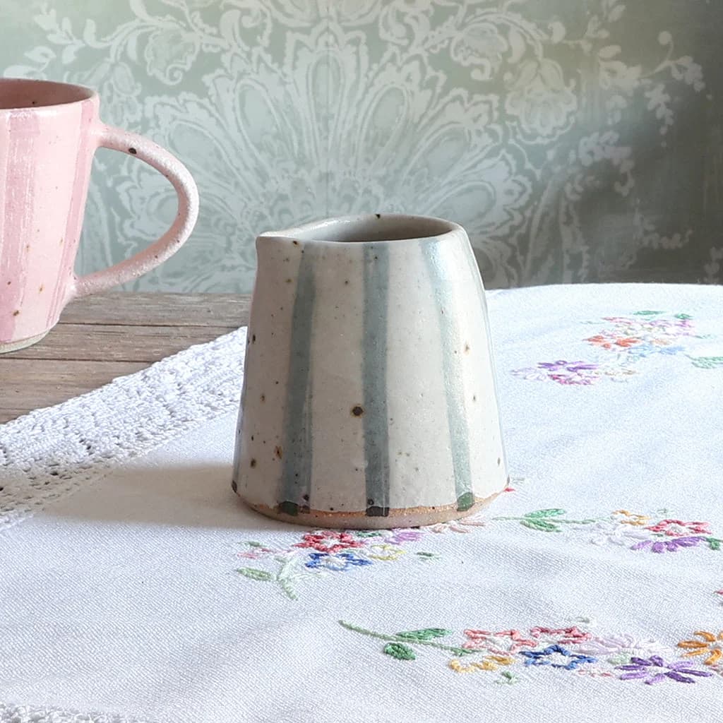 A handmade ceramic jug with a tapered shape, featuring a blue and white stripe design, set on a white embroidered tablecloth next to a striped pink mug.
