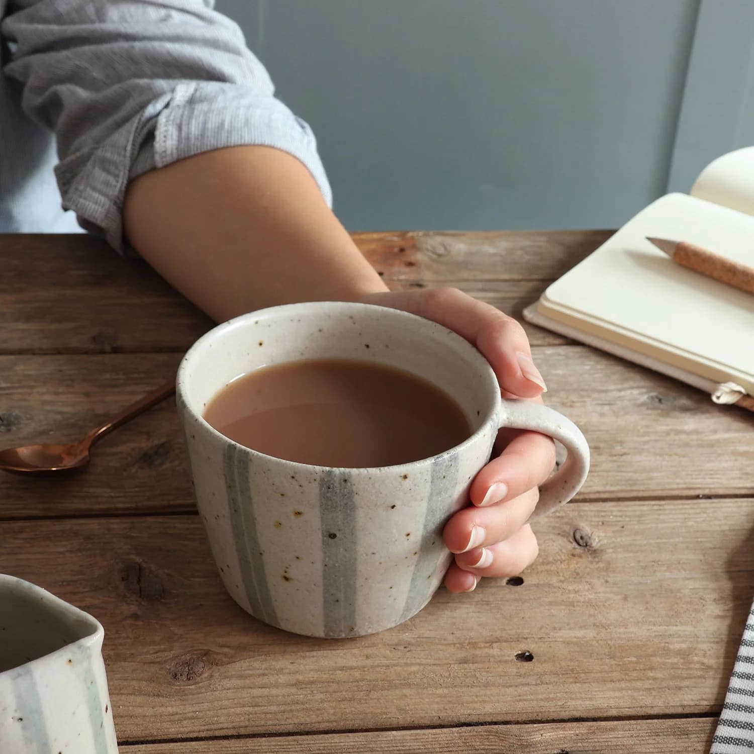A hand holding a blue and white stripe stoneware mug on a rustic wooden table with an open notebook and spoon nearby.