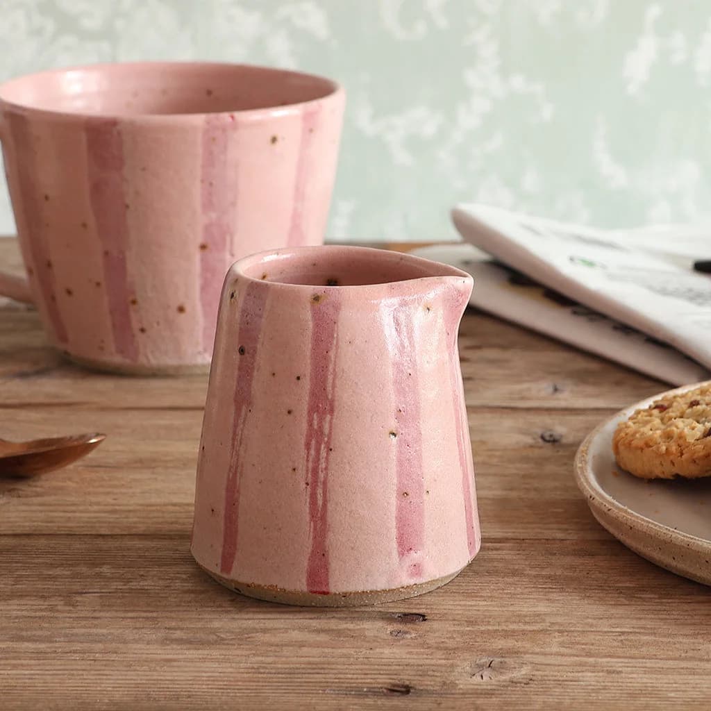 A pink stripy small handmade ceramic jug with a matching mug in the background, styled on a rustic wooden table with a plate of cookies and a newspaper.