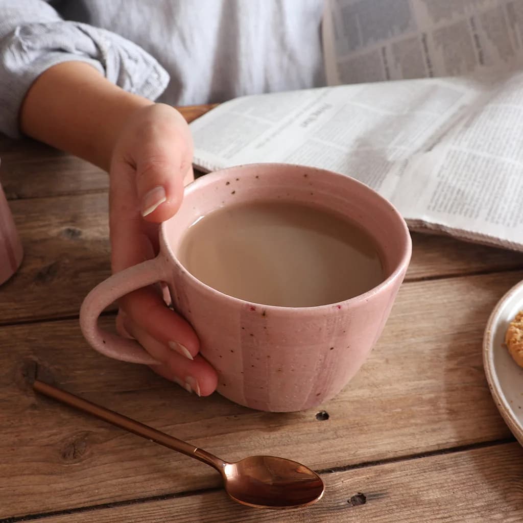 A person's hand holds a pink striped ceramic mug filled with tea on a rustic wooden table, with an open newspaper and a copper spoon nearby.