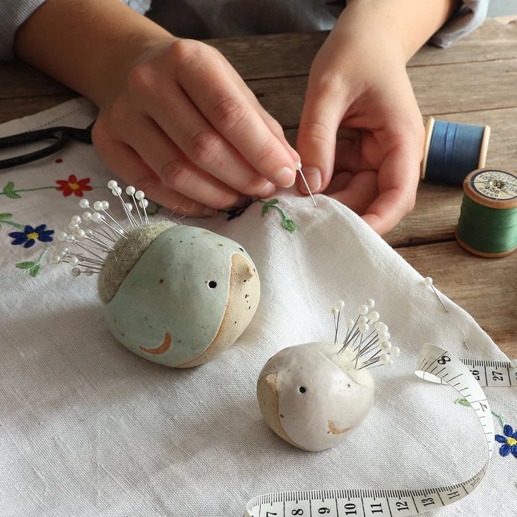 A large green ceramic and wool pin cushion bird holding pins sits on an embroidered cloth next to a small white pin cushion bird on a wooden surface. Hands can be seen poking a pin into the cloth behind the birds, next to two spools of thread and a sewing tape measure.