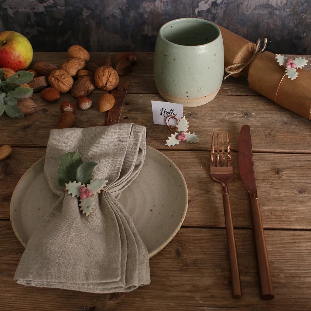 Set table with ceramic plates, green mug, and cutlery on a wooden surface. Napkin wrapped with ceramic napkin ring in the shape of a holly leaf.