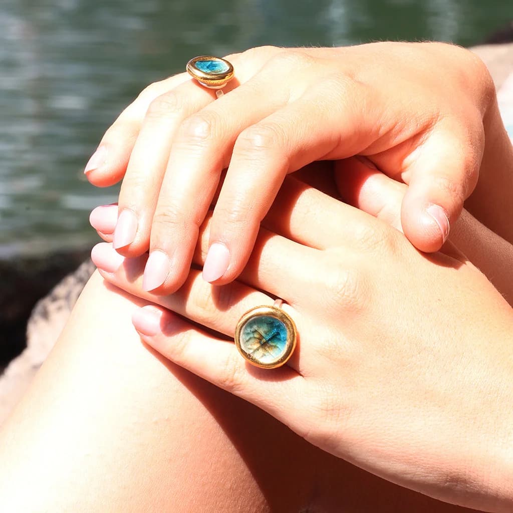 Two female hands wearing two sterling silver rings with ceramic, glass and gold charms. The glass is teal, white and brown.