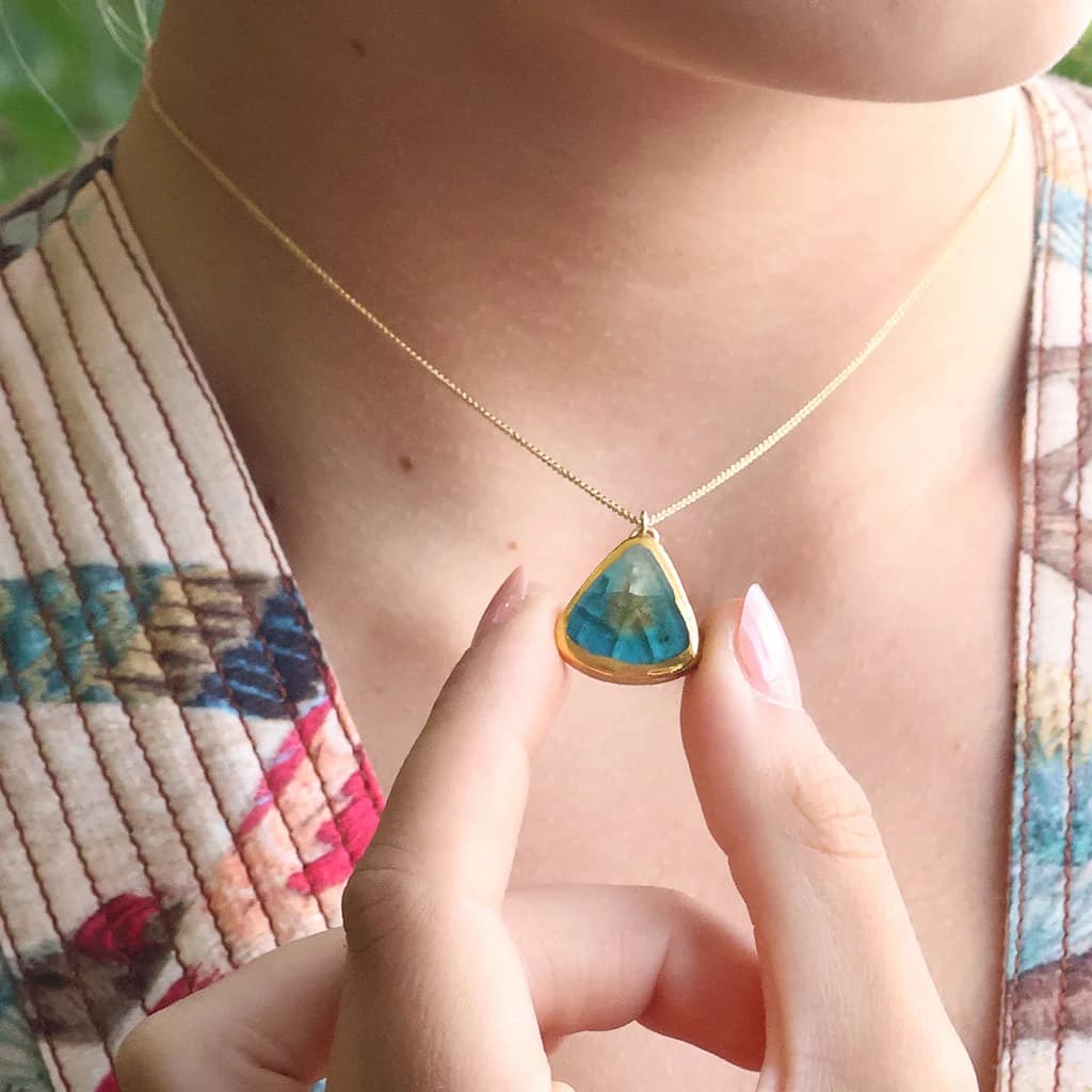 A women wears a delicate gold chain necklace with a teardrop-shaped pendant. The pendant is made from ceramic and glass in teal, white and brown with gold detailing, held gently between two fingers.