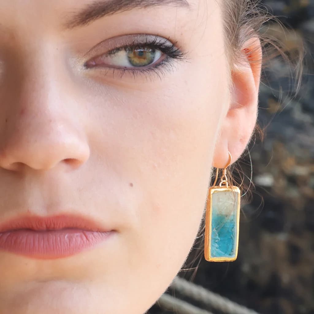 Close-up of a woman wearing a ceramic drop earring with teal and white glass with gold detailing.