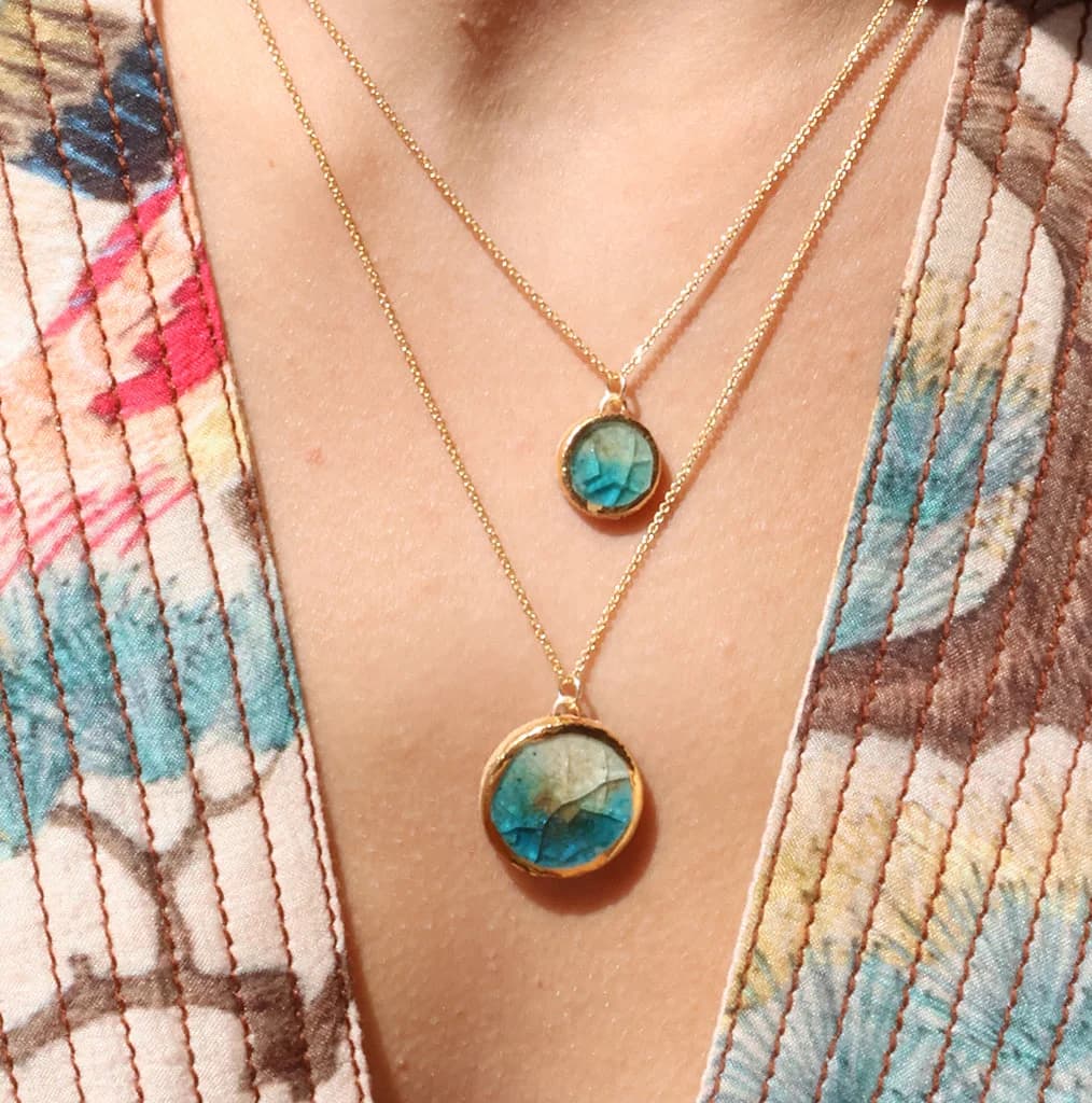 A women wearing two delicate gold chain necklaces, one with a small round pendant and one with a larger round pendant made from ceramic and teal coloured glass with gold detailing around the rim.