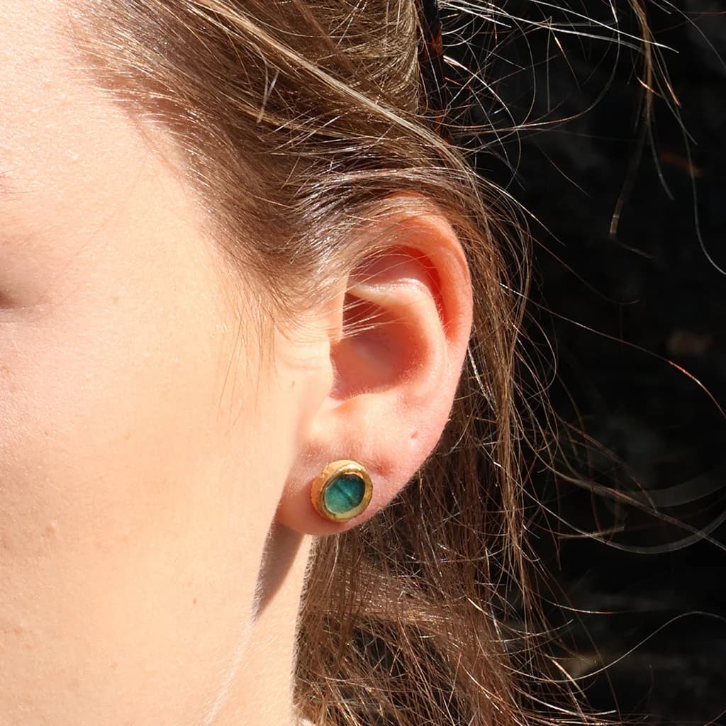 A women wearing a teal stud made from ceramic and glass with gold detailing, set against dark hair and a dark background.